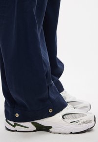 Pantalons amples bleu marine avec des accents de boutons aux poignets, associés à des chaussures de sport blanches dotées d'un mesh et de détails verts.