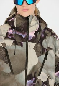 Mehrfarbige Jacke mit abstrakten Mustern in Grau, Lila und Pink. Das Material hat eine strukturierte Oberfläche mit Reißverschlussdetails und perforierten Akzenten.