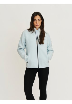 Vrouw die een lichtblauw fleecevest met rits draagt, met handen in de zakken, gecombineerd met zwarte leggings, staand tegen een effen achtergrond.