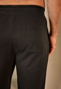 Pantaloni sport din bumbac negru cu un singur buzunar la spate, talie elastică și textură netedă. Focus pe o vedere apropiată a părții din spate.