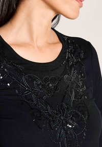 Schwarzes kurzärmliges Oberteil mit floralem Perlen- und Paillettenverzierung, Rundhalsausschnitt und glatter Stoffstruktur. Detaillierte Nähte betonen das Design.