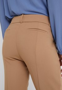 Pantalons beige ajustés en tissu lisse, dotés de deux poches arrière et d'une silhouette cintrée. Associés à un haut à manches longues bleu clair.