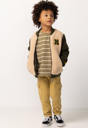 Kind met krullend haar, gekleed in een beige en groene fleecejack, gestreept t-shirt, beige cargobroek en zwart-witte sneakers, staat voor een witte achtergrond.