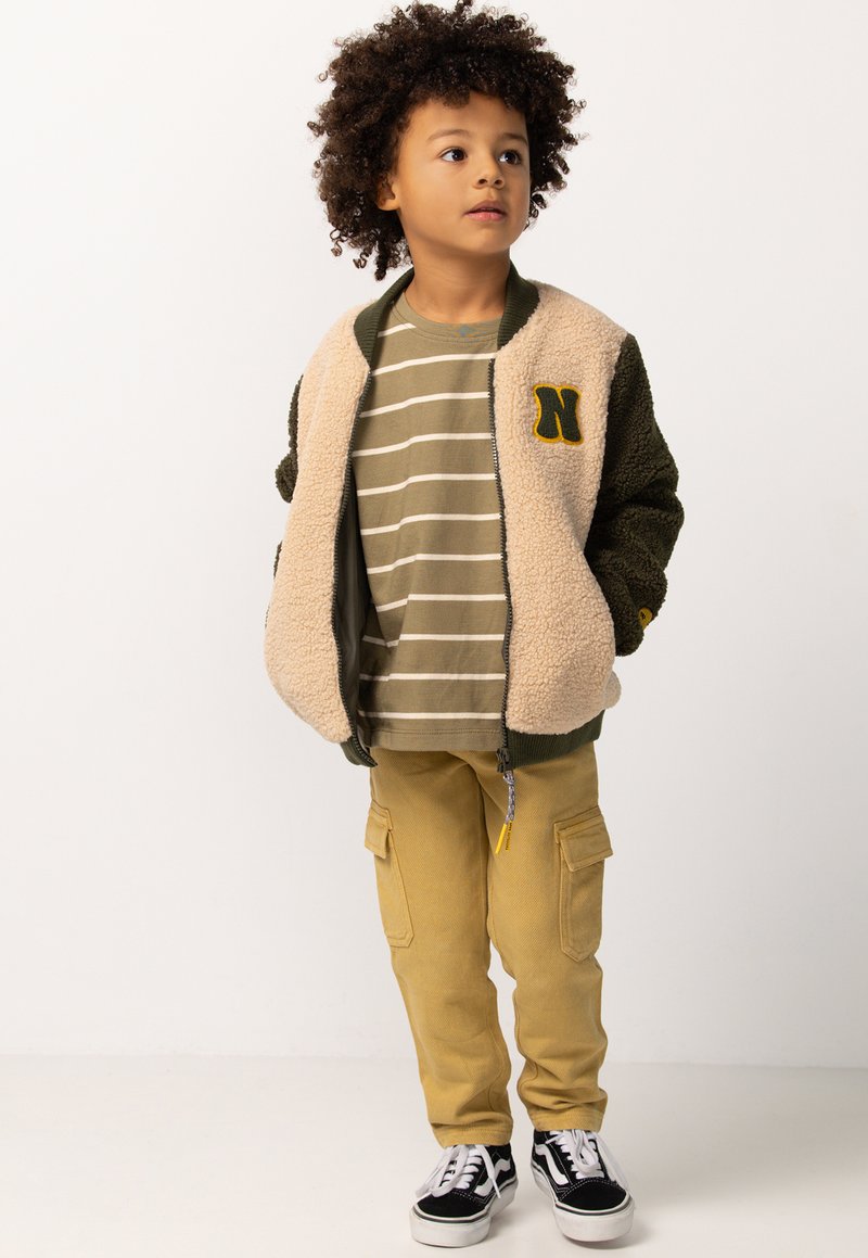 Kind met krullend haar, gekleed in een beige en groene fleecejack, gestreept t-shirt, beige cargobroek en zwart-witte sneakers, staat voor een witte achtergrond.