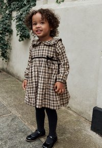 Brown plaid dress with a ruffled collar, black ribbon at the waist, black tights, and shiny black shoes, standing on a stone surface.