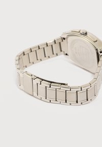 Ασημένιο βραχιόλι από μεταλλικούς κρίκους ενός ρολογιού χειρός με μερικώς ορατό εγχάρακτο κούμπωμα και την πίσω πλευρά της κάσας του ρολογιού σε απαλό φωτισμό.