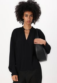 Blusa nera con scollo a V, davanti plissettato e maniche lunghe; presenta un anello con un piccolo fermaglio al colletto. Borsa in pelle nera in mano.