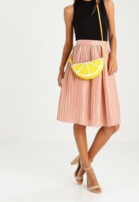 Żółta torebka typu crossbody w kształcie cytryny o teksturowanej powierzchni, noszona z czarną żebrowaną bluzką i różową plisowaną spódnicą midi, w połączeniu z cielistymi szpilkami.