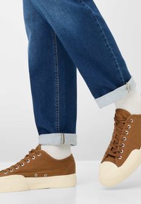 Sneakers in tela marrone con suole in gomma bianca, abbinate a jeans di denim blu risvoltati con un orlo azzurro chiaro.