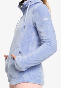 Vaaleansininen fleece-pusakka, joka on vetoketjullinen ja jossa on taskut, nyörikiinnitteinen huppu ja ribbipäät. Hihassa on hillitty logo. Pehmeä materiaali.