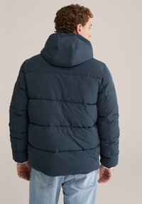 Man klädd i en mörkblå puffjacka med huva, vänd bort, i kombination med ljusblå jeans framför en enfärgad neutral bakgrund.