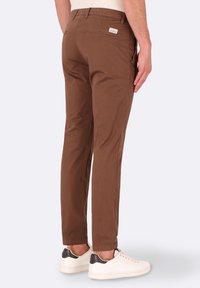 Chino marroni con vestibilità slim, dotati di tasche posteriori e di una piccola etichetta del marchio. Indossati con sneakers bianche, che mettono in risalto la texture liscia del tessuto.