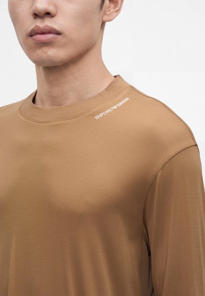 Крупен план на човек, носещ кафява риза с дълъг ръкав с отпечатано лого "EMPORIO ARMANI" близо до рамото на едноцветен фон.