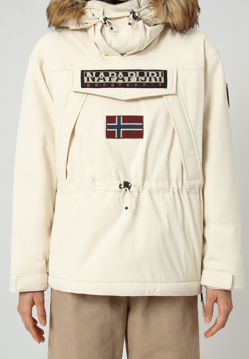 Cremefarvet pulloverjakke med hætte med pelskant, der har et stort logo-patch og et norsk flag-emblem, elastisk snøre i taljen.