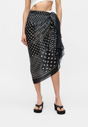 Kvinne som har på seg et svart-hvitt mønstret omslagskjørt og svarte platåsandaler, vist fra midje til føtter på hvit bakgrunn.