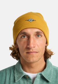 Cappello a coste giallo con logo a diamante e motivo a onda; indossato con una camicia in velluto a coste verde chiaro. Primo piano del viso e della parte superiore del corpo.