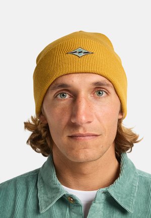 Żółta, prążkowana czapka z diamentowym logo i falistym motywem; noszona z jasnosz Shirt kordurowym. Zbliżenie na twarz i górną część ciała.