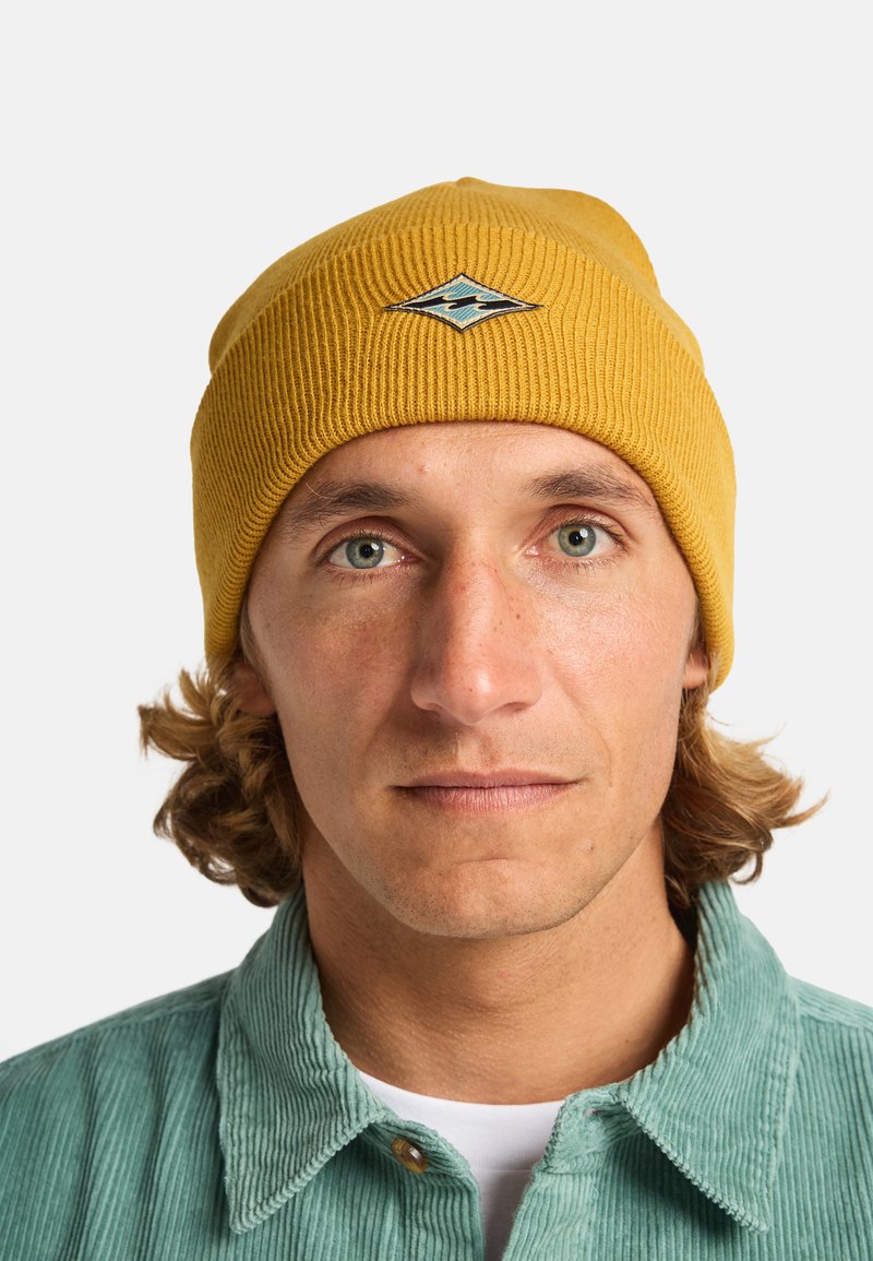 Cappello a coste giallo con logo a diamante e motivo a onda; indossato con una camicia in velluto a coste verde chiaro. Primo piano del viso e della parte superiore del corpo.