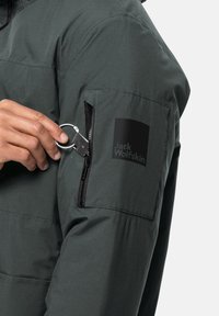 Dunkelgrüne gepolsterte Jacke mit einer Reißverschlusstasche am Ärmel, die ein Logo-Patch zeigt. Das Material wirkt glatt und strapazierfähig. Ein silberner Schlüsselring ist sichtbar.