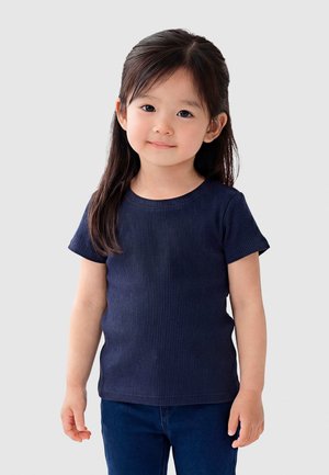 Niña joven con cabello largo y oscuro, vestida con una camiseta azul marino de canalé y pantalones oscuros, de pie contra un fondo gris claro.