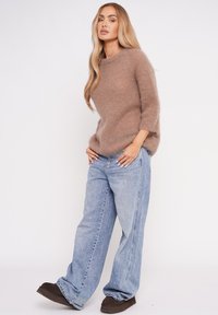 Frau, die einen flauschigen braunen Pullover, lockere hellblaue Jeans und dunkelbraune Schuhe trägt, steht vor einem weißen Hintergrund.