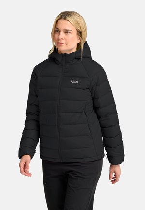 Vrouw draagt een zwarte Jack Wolfskin donzen jas met capuchon en ritssluiting aan de voorkant en zwarte broek, kijkt opzij tegen een effen achtergrond.