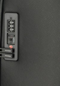 Superficie della valigia nera con texture, dotata di una combinazione di serratura, serratura approvata TSA e due etichette contrassegnate "GUESS" attaccate.