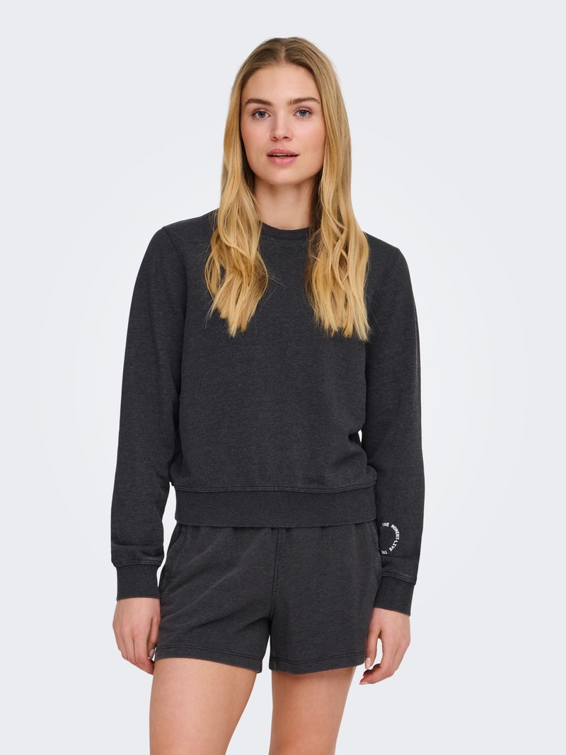 Jeune femme aux longs cheveux blonds, vêtue d'un sweat-shirt à manches longues gris foncé et d'un short assorti, sur un fond uni.