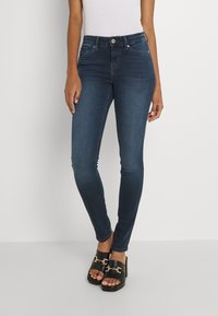 Smala mörkblå denimjeans med medelhög midja, fem-ficksdesign och knappar i guldnyans. Matchas med svarta platåsandaler.