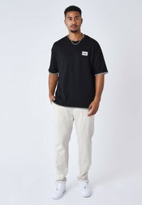 T-shirt nera oversize con cuciture bianche, caratterizzata da un piccolo logo patch, abbinata a pantaloni beige chiaro e sneakers bianche.