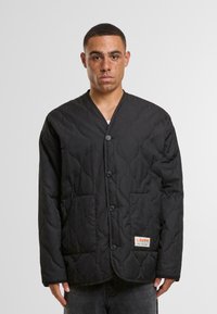 Black quilted jacket with a V-neck, button closure, two front pockets, and a branded label on the lower pocket. Smooth fabric texture.