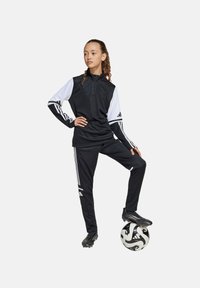 Survêtements de sport noir avec des manches blanches, trois bandes sur les bras et les pantalons, col zippé, design ajusté, associé à un ballon de soccer noir et blanc.