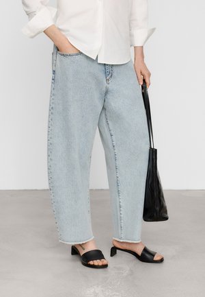 Persoon draagt lichtblauwe wijde jeans, wit overhemd, zwarte sandalen met hak en houdt een zwarte leren tote bag vast.