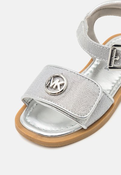Sandales argentées avec une sangle métallique texturée, un accent de logo circulaire et une semelle plate marron. Fermeture à boucle sur la sangle arrière réglable.