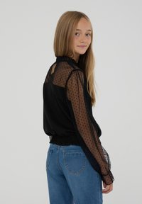 Blusa nera con maniche trasparenti a pois, scollatura arricciata e vita elastica. Indossata con jeans azzurri chiari, caratterizzati da una vestibilità comoda.