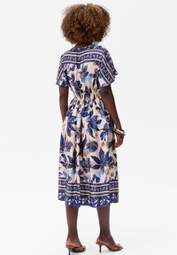 Florale jurk met korte mouwen, voorzien van een getailleerde taille en een knielange zoom. Kleuren zijn crème, blauw en goud met decoratieve patronen.