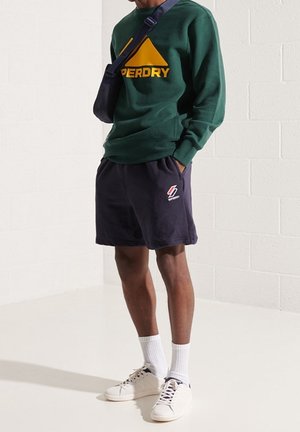 Zelená mikina se žlutým logem „Superdry“, námořnicky modré šortky, bílé ponožky a bílé tenisky. Součástí outfitu je kabelka přes rameno.