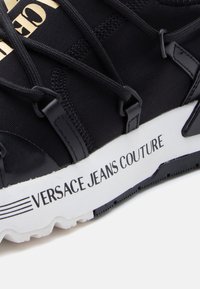Μαύρο αθλητικό παπούτσι με υφαντό ύφασμα στην επάνω επιφάνεια, ελαστικά κορδόνια και λευκή σόλα με την επιγραφή "VERSACE JEANS COUTURE" σε μαύρο χρώμα.