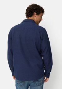 Man with curly hair wearing a long-sleeve navy blue shirt and blue jeans, facing away against a plain white background.