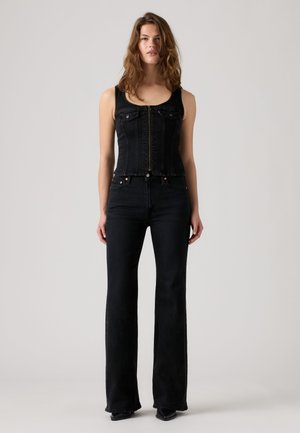 Kvinde med bølget brunt hår iført en sort ærmeløs denimvest med lynlås foran og sorte udvidede jeans, stående mod en ensartet lys baggrund.