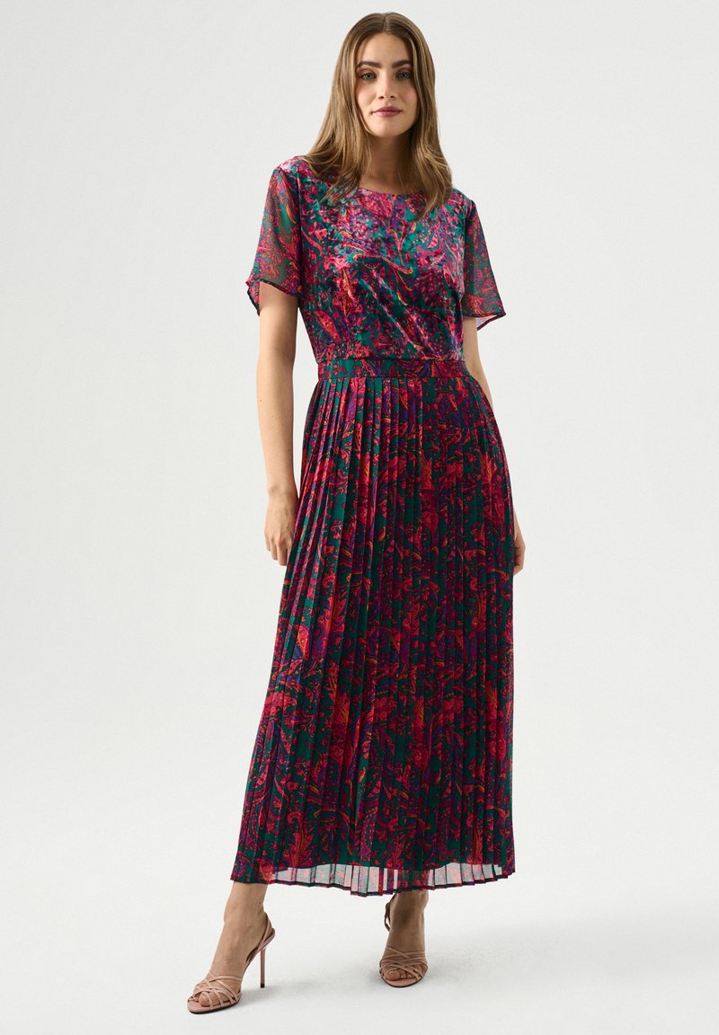 Plissiertes Maxikleid mit einem floralen Muster in rosa und grünen Tönen, kurzen Ärmeln und einer fließenden Silhouette. Tragen mit nude-farbenen Riemchenschuhen.
