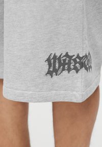 Ανοιχτό γκρι υφασμάτινες σορτσάκια με μαύρο αφηρημένο γραφικό κοντά στο τελείωμα, φορεμένα από ένα άτομο με ορατά κάτω πόδια.