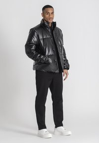 Giacca a vento nera e lucida con colletto alto, chiusura a zip e tasche laterali, abbinata a pantaloni neri e sneakers bianche.