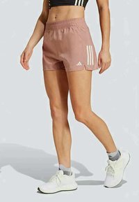 Egy nő mauve színű Adidas sportnadrágot visel, fehér csíkkal és logóval, fehér sportcipővel és szürke bokazoknival párosítva, egy egyszínű háttér előtt állva.