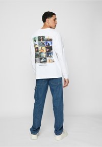 Hvid langærmet skjorte med et collage af grafiske billeder på ryggen, med tekst, parret med blå jeans og hvide sneakers.