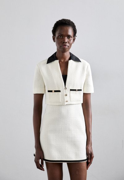 White tweed cropped jacket with black collar, front button closure, and two pocket accents; paired with a form-fitting white tweed mini skirt.