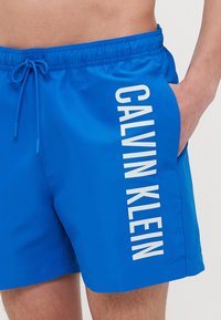 Blaue Badeshorts aus glattem Stoff mit einem elastischen Bund und verstellbarem Kordelzug sowie einem weißen, vertikal auf der Seite aufgedruckten Logo.