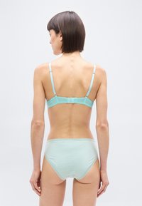 Γυναίκα εμφανίζεται από πίσω φορώντας ανοιχτό μπλε σουτιέν και εσώρουχα ψηλόμεσα μπροστά σε απλό φωτεινό φόντο.