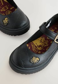 Chaussures Mary Jane noires avec un dessus en cuir texturé, semelle plate, quincaillerie dorée et semelle intérieure bordeaux ornée d'un motif fantaisiste.