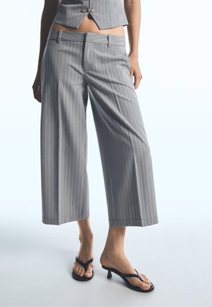 Femme portant un pantalon large gris à rayures raccourci, un gilet assorti et des sandales noires à talons type tongs, debout sur un fond blanc.